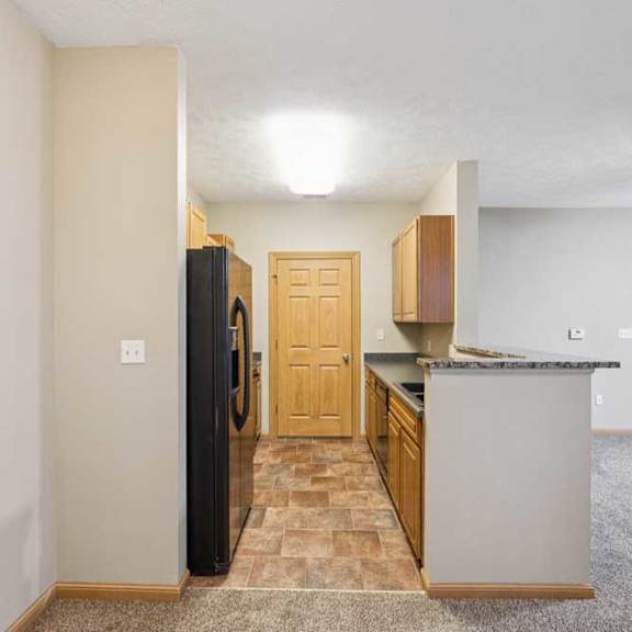 Thumbnail 4 of 20 Galley-style apartment kitchen with wood cabinets, black refrigerator, laminate countertops, and tile flooring, opening to a carpeted living area.
