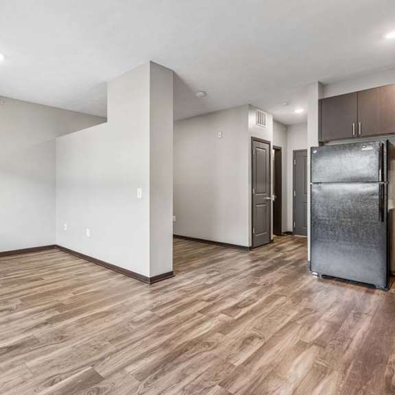 Thumbnail 6 of 15 Spacious studio apartment with wood-style flooring, recessed lighting, an open layout, and a compact kitchen with black appliances.