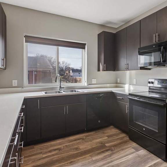Thumbnail 20 of 34 Modern apartment kitchen with dark wood cabinetry, black appliances, white countertops, and a window over the sink.