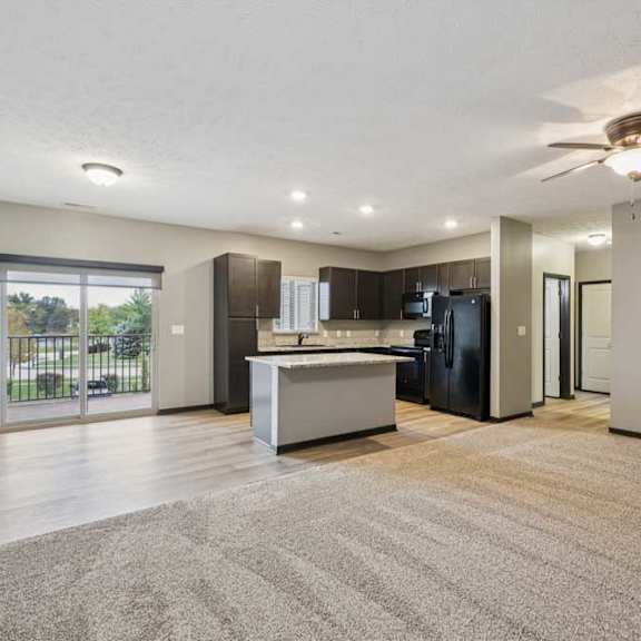 Thumbnail 7 of 33 A spacious, carpeted living room open to a modern kitchen with hardwood flooring and dark wooden cabients.