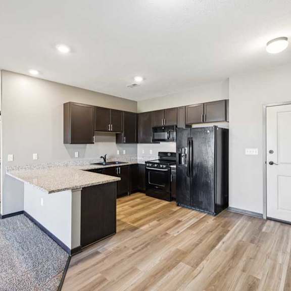 Thumbnail 15 of 30 Apartment kitchen with L-shaped counter, dark wood cabinets, black refrigerator and oven, and adjacent entry and storage doors.