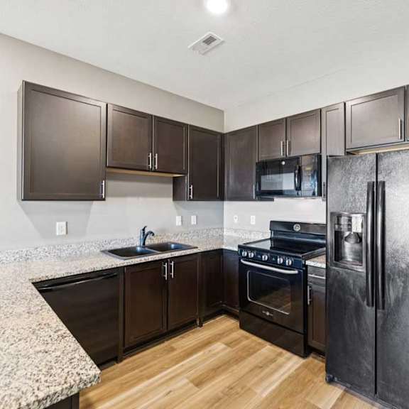Thumbnail 16 of 30 Modern apartment kitchen with granite-look countertops, dark cabinetry, black oven and refrigerator, and wood-style flooring.