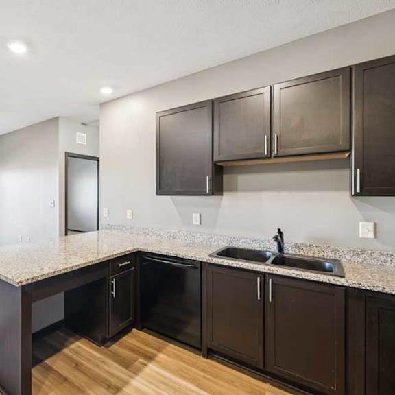 Thumbnail 17 of 30 Kitchen with extended breakfast bar, double sink, dark cabinets, and open view into the carpeted living room and balcony.
