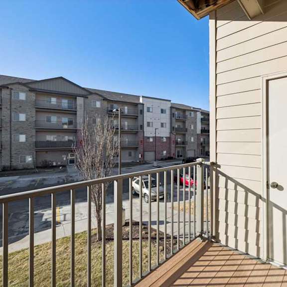 Thumbnail 9 of 23 A balcony with a white door and a metal railing overlooks a parking lot and apartment buildings.