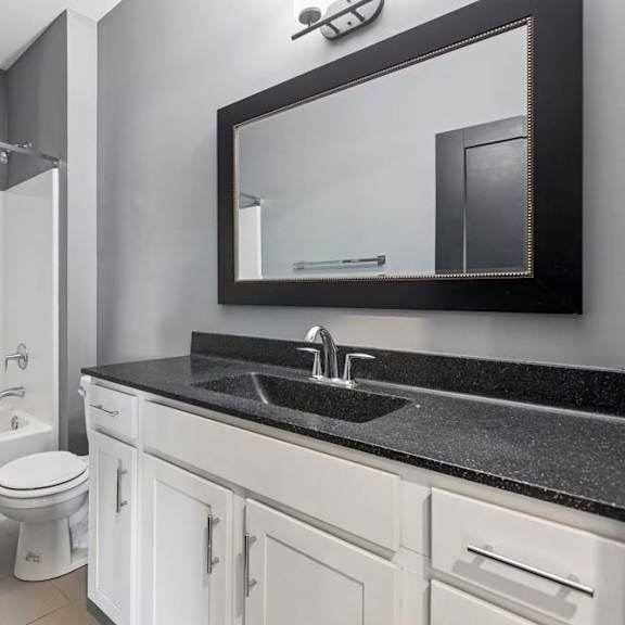 Thumbnail 34 of 46 Black-framed mirror and speckled dark countertop contrast with a white vanity and tub-shower combo in this apartment bathroom.
