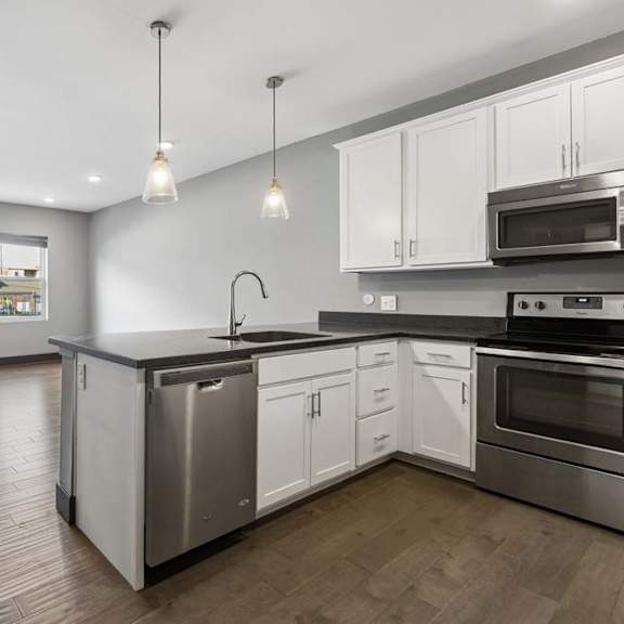 Thumbnail 27 of 46 Open-concept apartment kitchen flows into a sunlit living area, with white cabinetry, dark counters, pendant lights, and stainless steel appliances.