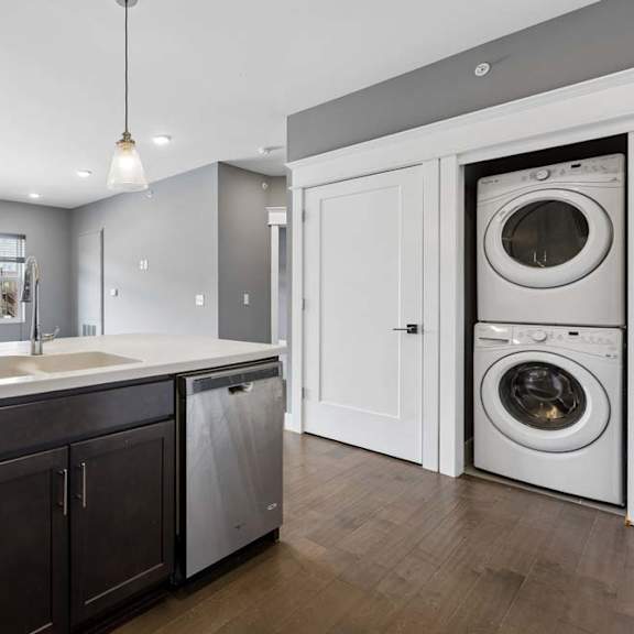 Thumbnail 38 of 46 Stacked in-home laundry sits just off the kitchen, beside a dark cabinet island with a deep sink, dishwasher, and pendant lights.