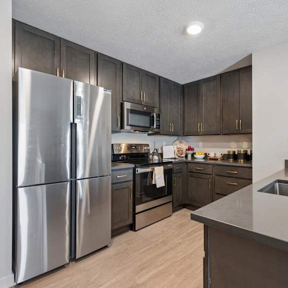 Thumbnail 26 of 39 Modern apartment kitchen with dark wood cabinets, stainless steel appliances, light wood flooring, and a large island with sink and countertop seating area.