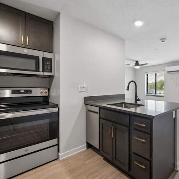 Thumbnail 43 of 52 Open-concept apartment kitchen with dark cabinetry, stainless steel appliances, a center island with sink, and a spacious living area with a glass patio door.