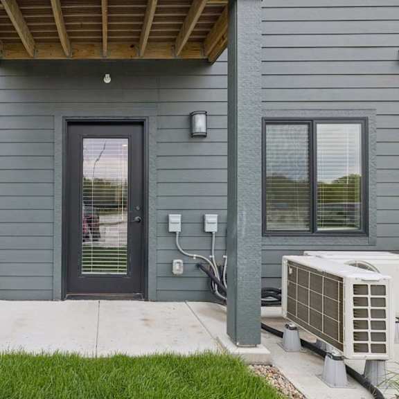 Thumbnail 52 of 52 Covered ground-floor apartment patio with a concrete surface, glass-paneled entry door, exterior wall light, and landscaped surroundings.