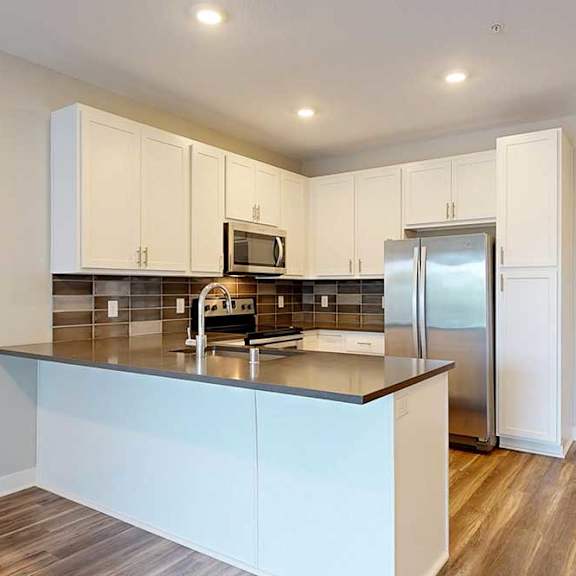 Thumbnail 5 of 25 A kitchen with white wooded cabinets, quartz countertops, a peninsula, stainless steel appliances, and a designer backsplash.
