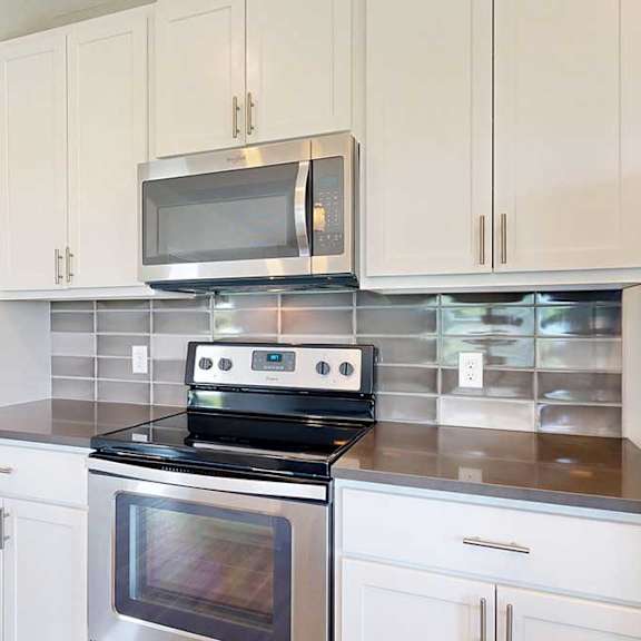 Thumbnail 5 of 29 A close up on a stainless steel range and microwave in a kitchen with white wooden cabinets, quartz countertops, and a designer tile backsplash.