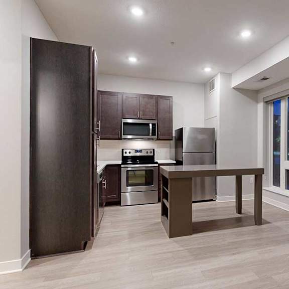 Thumbnail 14 of 21 A living area with hardwood-style flooring and kitchen with dark wooden cabinets.