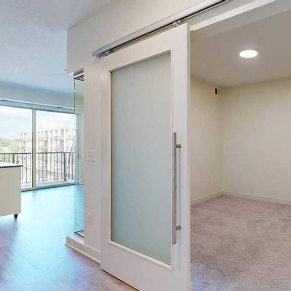 Thumbnail 3 of 17 A bright room features a frosted glass sliding door, beige carpeting, and modern wood flooring. A large window offers a view of a balcony and buildings.