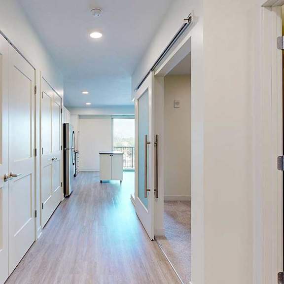 Thumbnail 2 of 17 Modern, bright apartment hallway with wood flooring, white walls, and multiple doors. An open kitchen with stainless steel appliances is visible ahead.