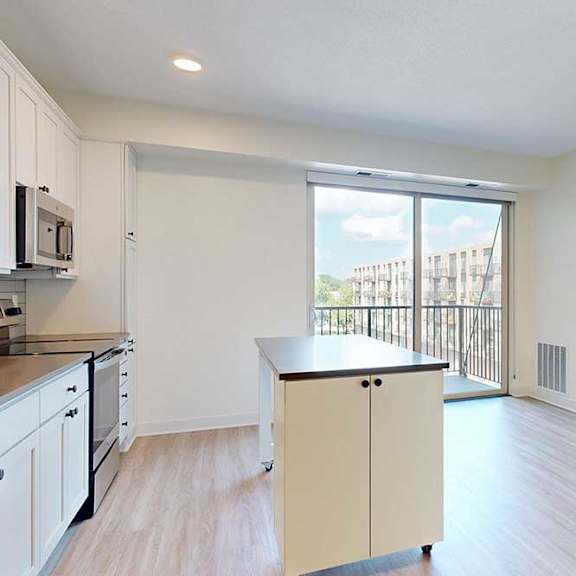 Thumbnail 8 of 17 A modern, bright kitchen with white cabinets, stainless steel appliances, and a small island. A large window with a view of a building outside adds natural light.