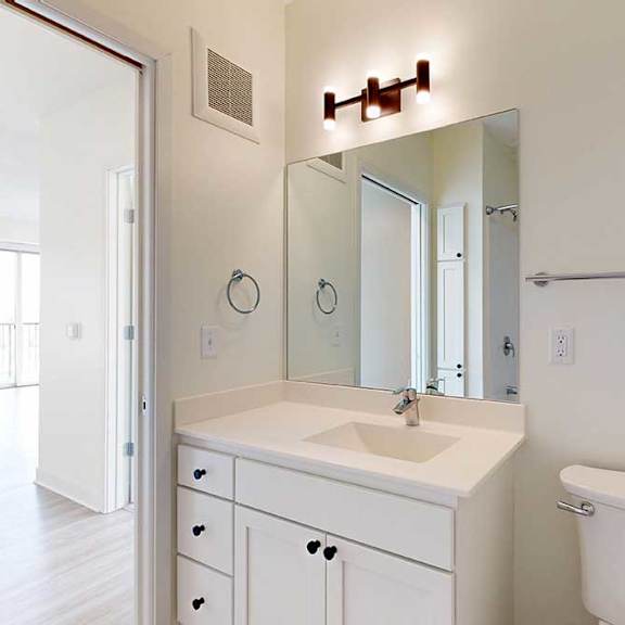 Thumbnail 5 of 23 Modern bathroom featuring a white vanity, large mirror, chrome fixtures, and a toilet. Natural light streams in from the adjacent room.