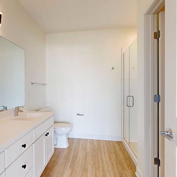 Thumbnail 15 of 23 Modern bathroom with white cabinets, a large mirror, single sink, toilet, glass shower enclosure, and wood-look flooring.