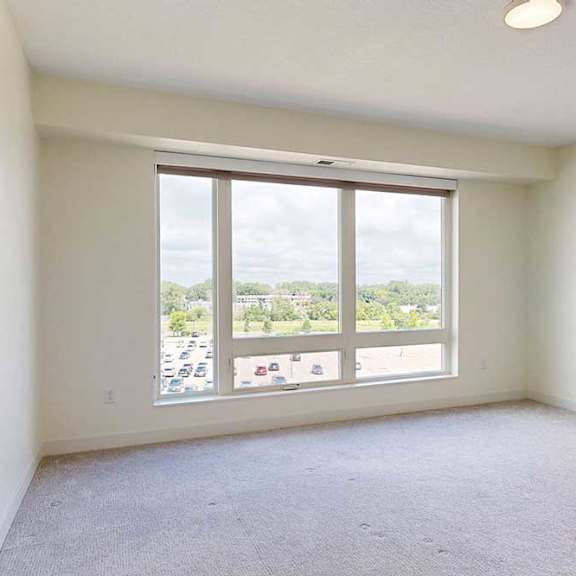 Thumbnail 13 of 23 Bright, empty beroom with light-colored walls and carpet, featuring a large window that offers a view of greenery and parking below.