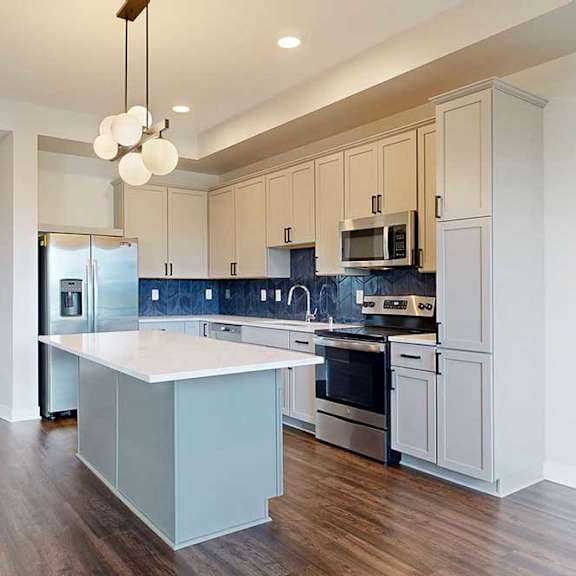 Thumbnail 7 of 31 Modern kitchen with white cabinets and a blue backsplash, featuring stainless steel appliances. A central island with a white countertop is illuminated by a stylish pendant light, and dark wooden floors add warmth.
