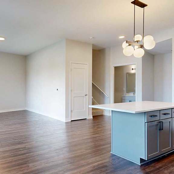 Thumbnail 10 of 31 Modern kitchen and living area in neutral tones. Wood flooring, white walls, pendant light fixture, and open island countertop create a spacious feel.