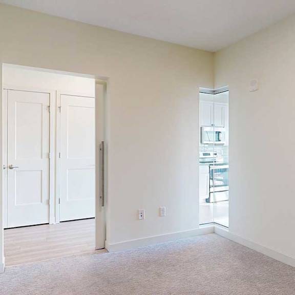 Thumbnail 5 of 19 Empty room with beige walls and carpet, featuring a white door leading to another space with wooden floors. A glimpse of a modern kitchen is visible.
