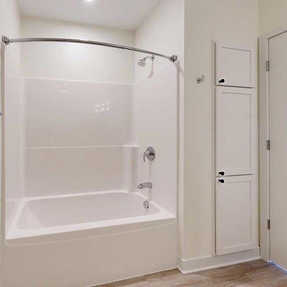 Thumbnail 6 of 26 Simple bathroom with a white bathtub-shower combo, curved shower rod, and beige walls. Wooden floor, towel bar, and a cabinet beside a door.