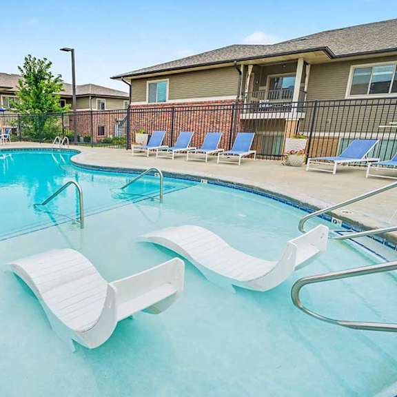 resort-style apartment pool with curved design, shallow sun shelf and in-water lounge chairs, surrounded by deck seating and fenced residential buildings.