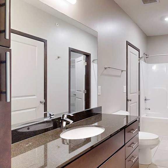 Thumbnail 23 of 25 An attached bathroom with a granite sink vanity, dark wooden cabinets, and a tub shower.