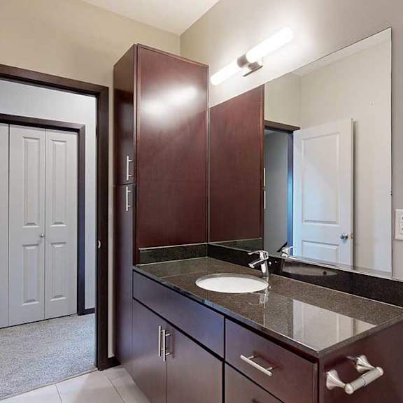 Thumbnail 22 of 25 A bathroom with a granite vanity sink, dark wooden cabinets, a tall storage cabinet, and an oversized mirror.