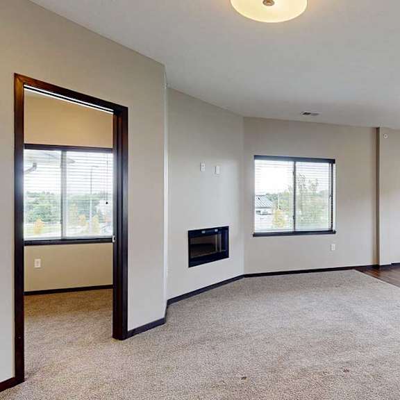 Thumbnail 7 of 25 A carpeted living room with several windows, an electric fireplace, and an open door to a den space.