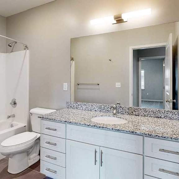 Thumbnail 25 of 26 A bathroom with a granite vanity, white wooden cabinets, an oversized mirror with overhead lighting, and a tub shower.