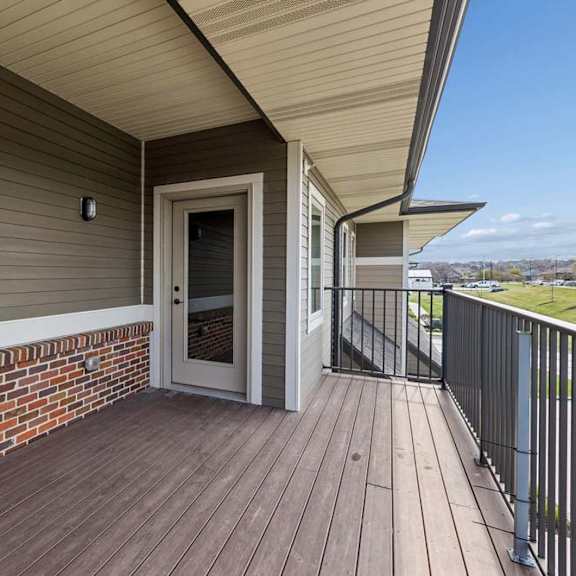 Thumbnail 10 of 18 Covered private balcony with wood-look decking, brick accents, and direct access to the apartment entrance.