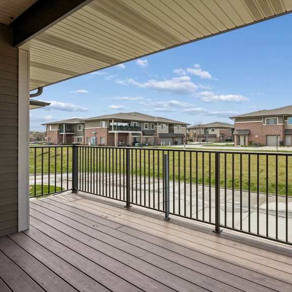 Thumbnail 9 of 18 Covered apartment balcony with wood-look decking, black railing, and views of nearby community buildings.