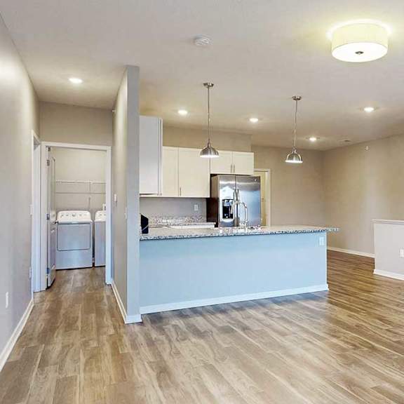 Thumbnail 5 of 26 A living room with hardwood-style flooring, open to a kitchen with white wooden cabinets.