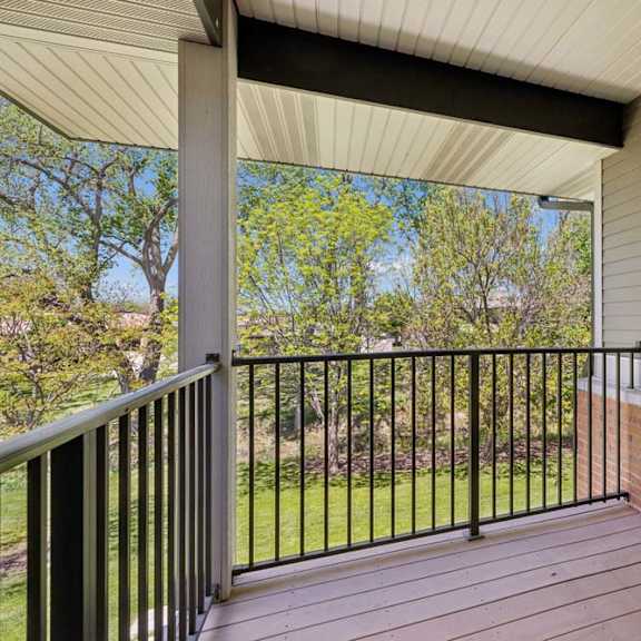 Thumbnail 21 of 30 Private apartment balcony with black railing, wood-look decking, and a leafy green courtyard view.
