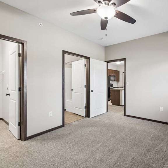 Thumbnail 3 of 19 Empty carpeted bedroom with ceiling fan, window with blinds, and doorways to bathroom and kitchen area.
