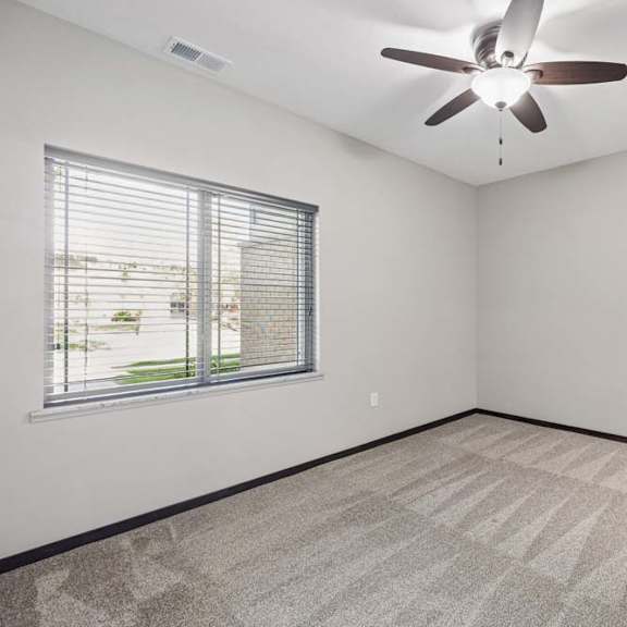 Thumbnail 7 of 19 Bright carpeted bedroom with ceiling fan, neutral walls, large window with blinds, and dark trim.