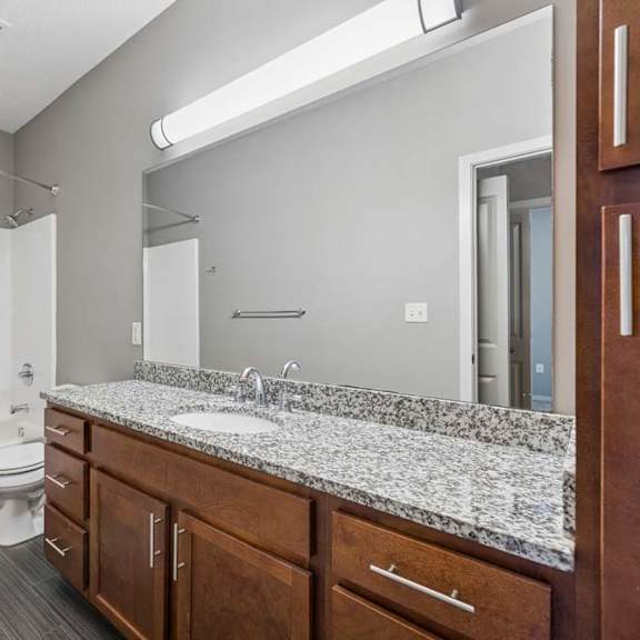 Thumbnail 14 of 16 Apartment bathroom with tub and shower combo, speckled countertop, wood vanity, large mirror, and gray tile floor.