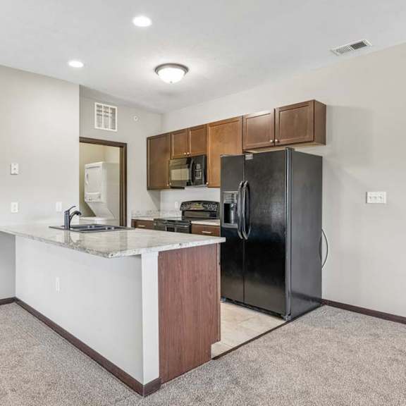 Thumbnail 39 of 48 Open apartment kitchen with granite-style island, dark cabinets, black appliances, tile flooring, and carpeted living area.