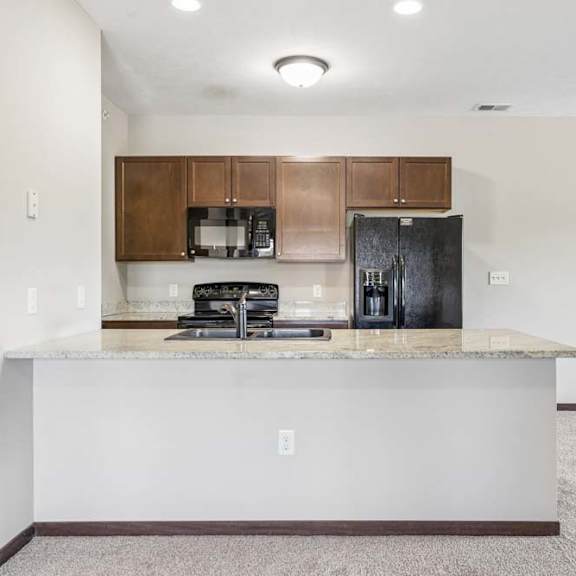 Thumbnail 35 of 48 Open apartment kitchen with granite-style breakfast bar, dark cabinets, black appliances, and carpeted living area.