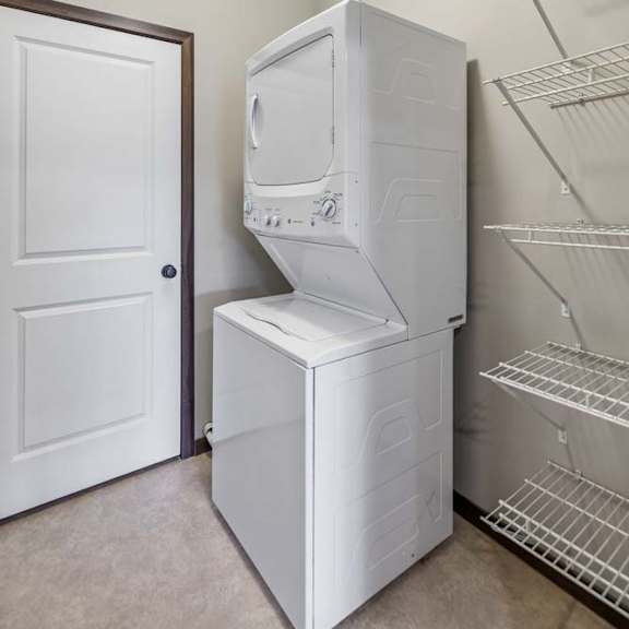 Thumbnail 31 of 48 Apartment laundry room with stacked washer and dryer, wire shelving, overhead light, and neutral walls.