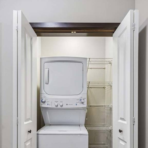 Thumbnail 23 of 59 Apartment laundry closet with stacked washer and dryer, wire shelving, and double doors.
