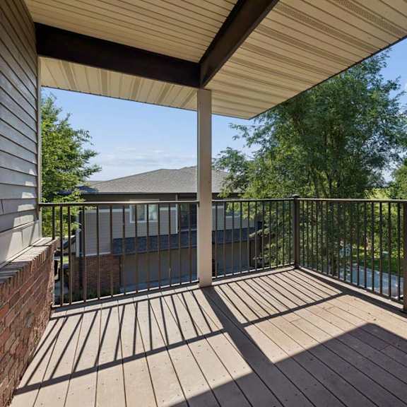Thumbnail 15 of 16 Covered apartment balcony with wood-look decking, black metal railings, and views of mature trees and landscaped green space.