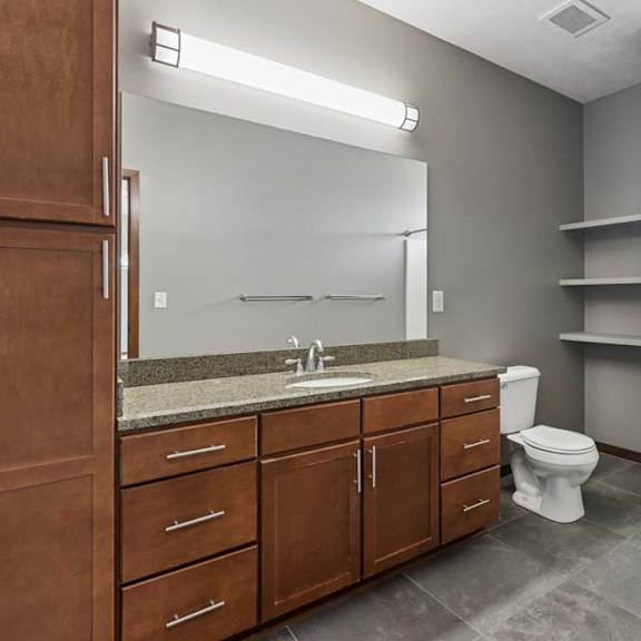 Thumbnail 13 of 16 Apartment bathroom with wood cabinetry, stone-look vanity counter, open shelving, gray tile flooring, and a walk-in shower.