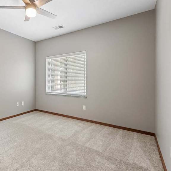 Thumbnail 8 of 16 Bright, empty apartment bedroom with neutral carpet, gray walls, a ceiling fan, and a large window with blinds.
