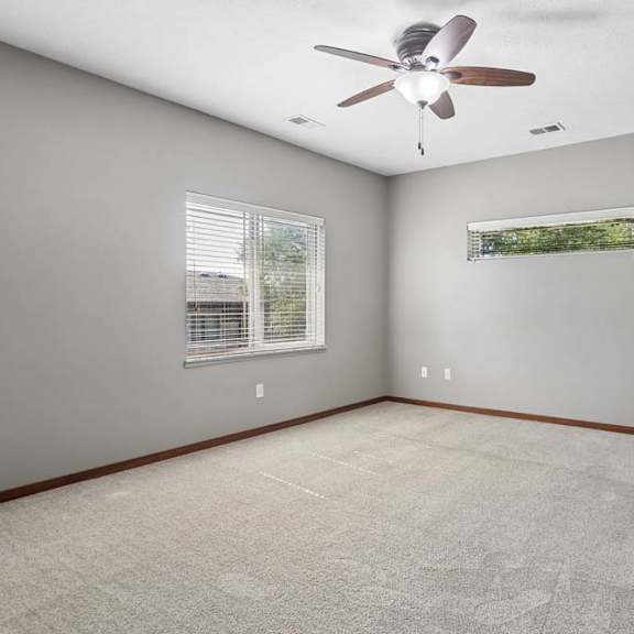 Thumbnail 10 of 16 Bright, empty apartment bedroom with neutral carpet, gray walls, ceiling fan, and two windows with blinds.