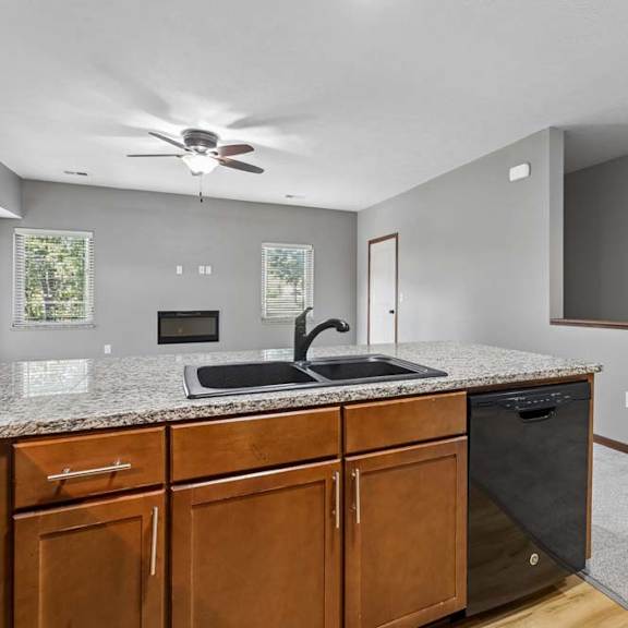 Thumbnail 4 of 16 Granite kitchen peninsula with double sink overlooking a bright living space with fireplace, ceiling fan, and stairway access.