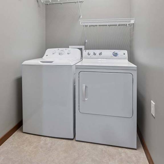 Thumbnail 14 of 16 In-home apartment laundry room with a white washer and dryer, wall-mounted wire shelving, and neutral gray walls.