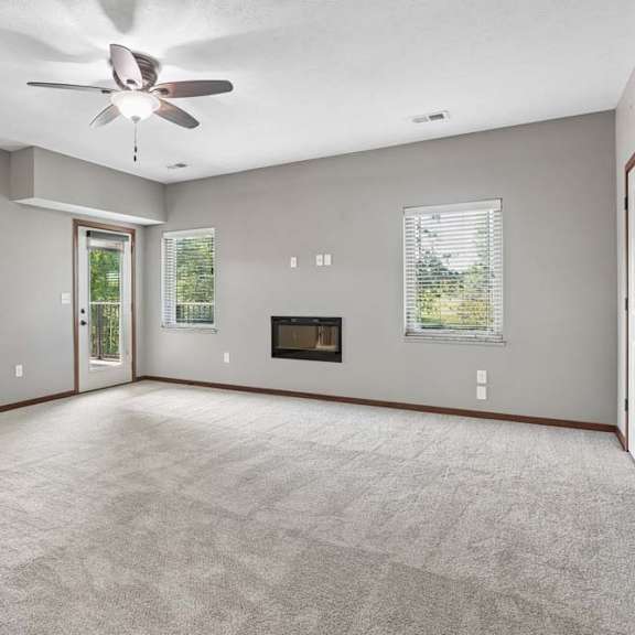 Thumbnail 6 of 16 Bright, empty apartment living room with carpet, ceiling fan, two windows, a fireplace, and a glass door to the balcony.